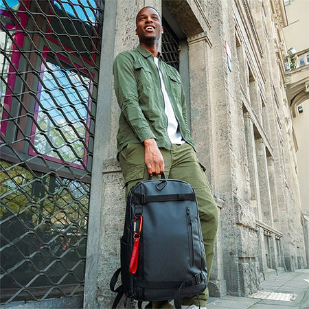 Herren Rucksack in schwarz von sons of aloha AHI Mann ist gegen eine Hauswand gelehnt und traegt ein gruenes Hemd und eine gruene Hose