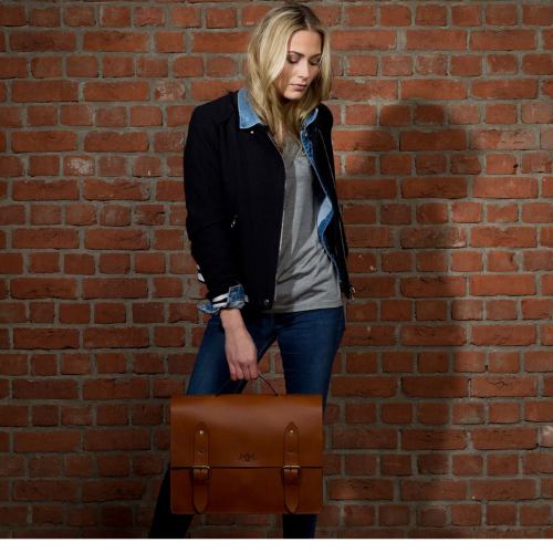 Frau mit brauner Ledertasche in einer Hand, vor einer Ziegelsteinmauer