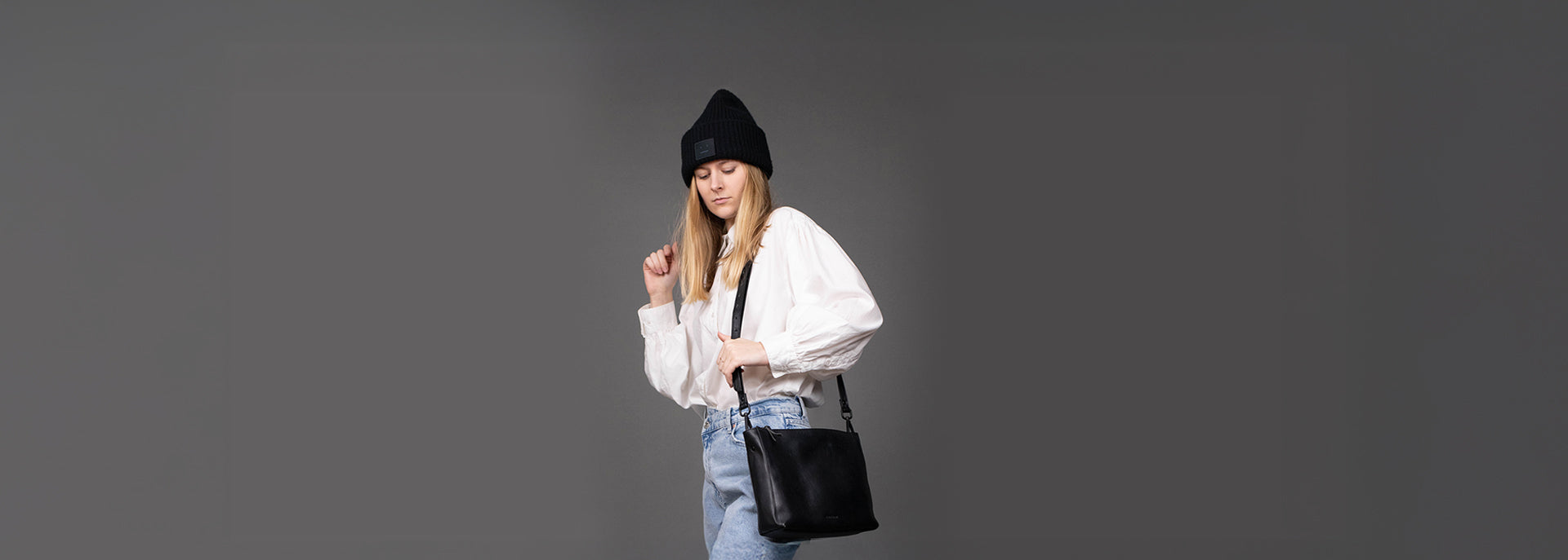 Studiobild grauer Hintergrund mit weiblichen Model mit schwarzer Ledertasche, schwarzer Beanie, weißen Hemd und heller Jeans