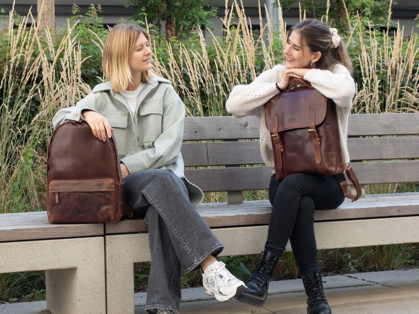 Zwei Frauen mit braunen Lederrucksäcken, sitzend auf einer Parkbank