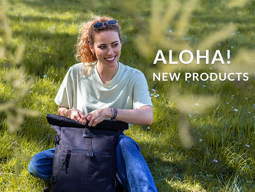 Frau sitzend auf einer Wiese mit Rolltop Rucksack und dem Text "Aloha! New Products" als Header