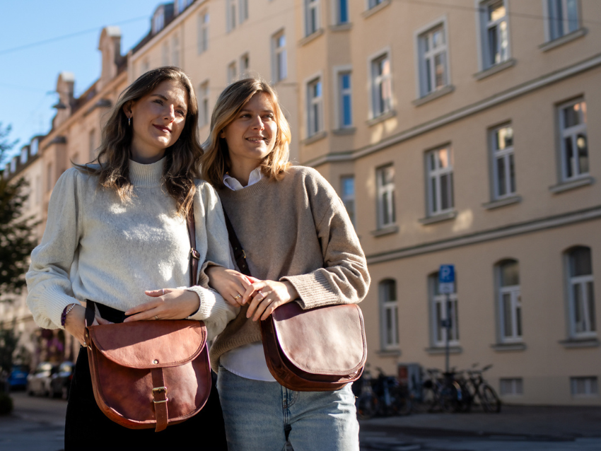 Zwei Frauen in einer Innenstadt mit jeweils einer braunen Umhängetasche