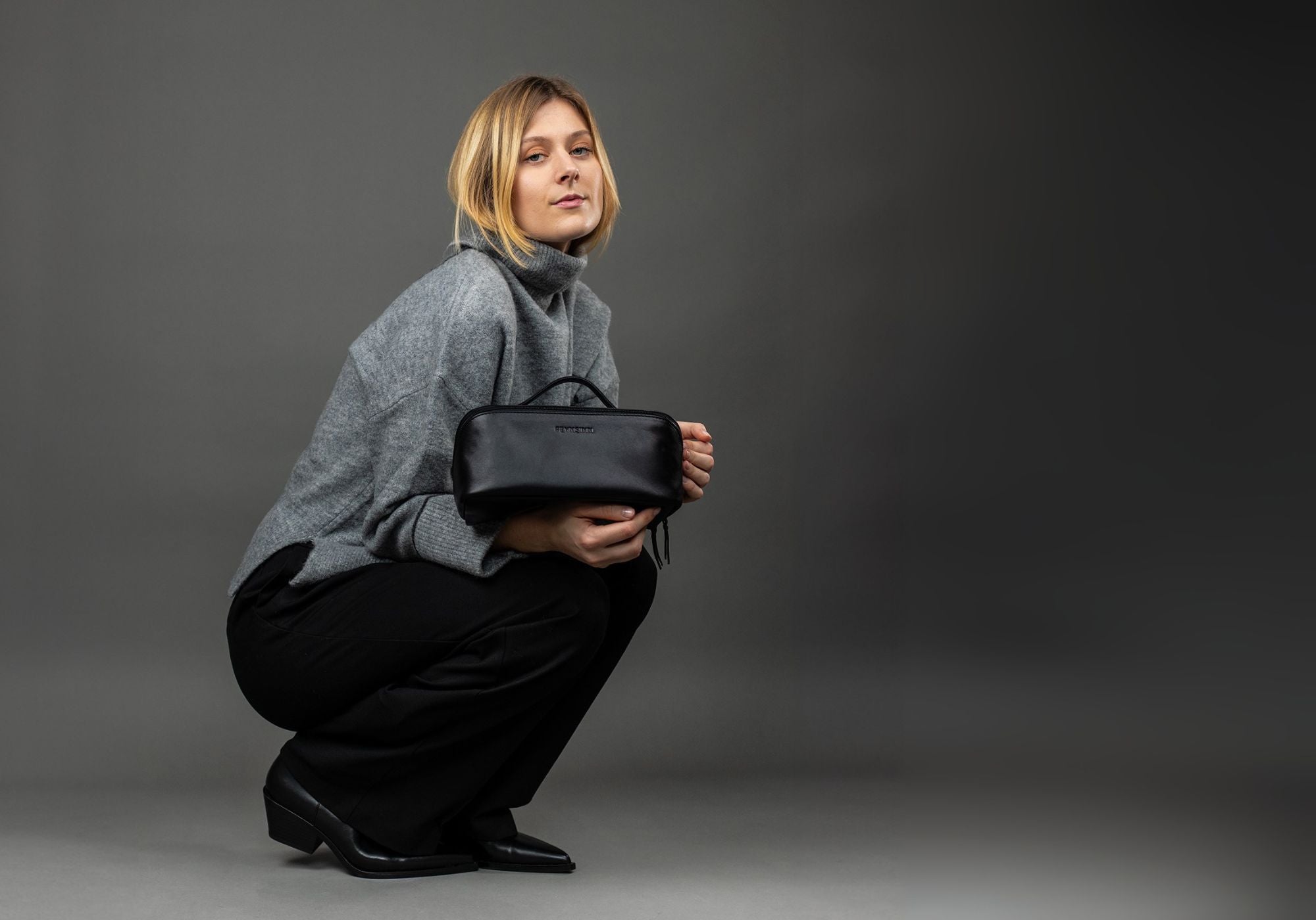 FEYNSINN Kulturbeutel Studioaufnahme mit Waschbeutel Leder schwarz BO Model traegt einen grauen Pulli und eine schwarze HOse