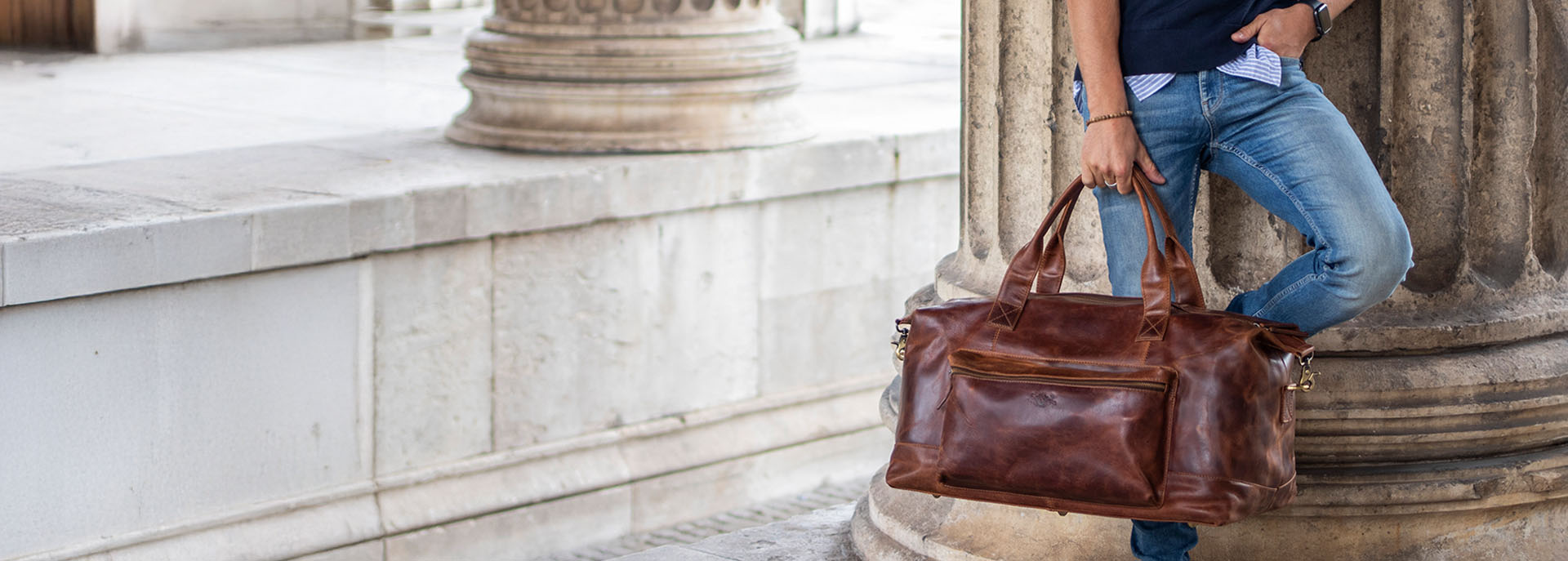 Mann lehnt an Säule mit brauner Leder Reisetasche in der Hand. Die Tasche hat eine schöne Patina
