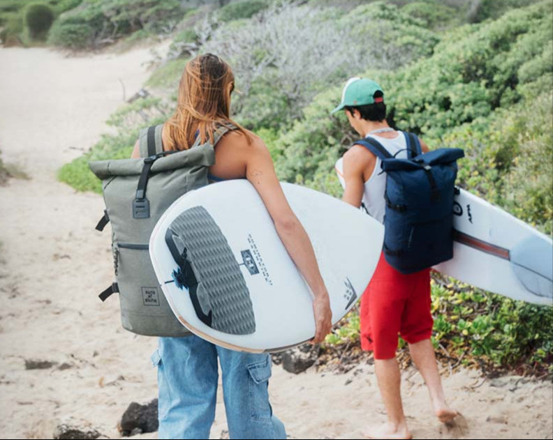 Mann und Frau mit Rucksäcken in oliv und blau mit Surfbrettern unterwegs zum Meer