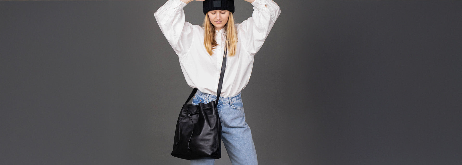 Model mit schwarzer Beuteltasche aus Glattleder, Hemd und Jeans im Studio mit grauen Hintergrund