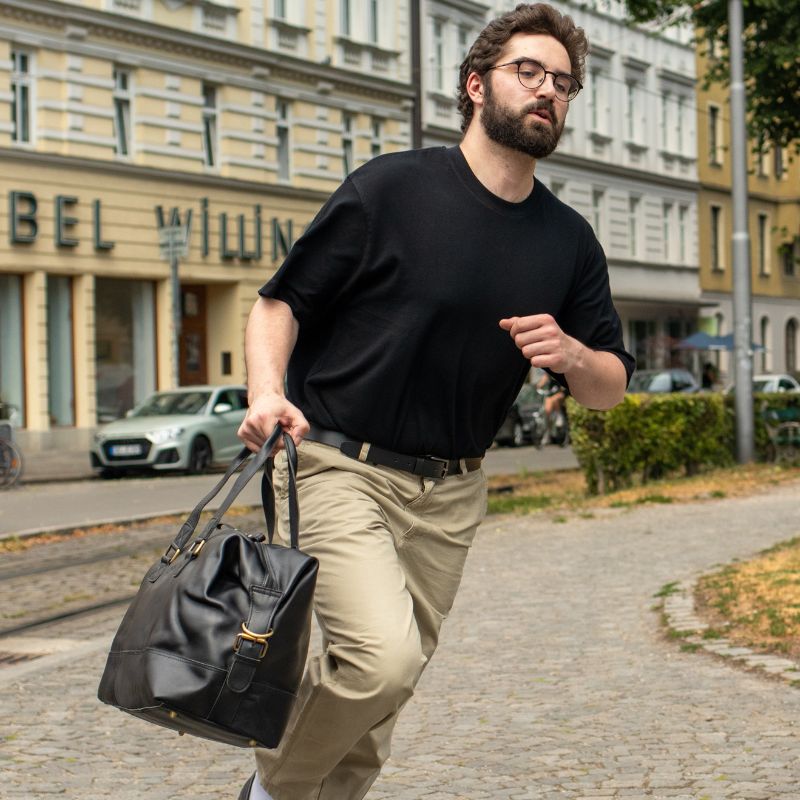 Mann rennt in der Stadt und traegt eine Reisetasche Leder in schwarz und hat ein schwarzes T-Shirt und eine beige Hose an